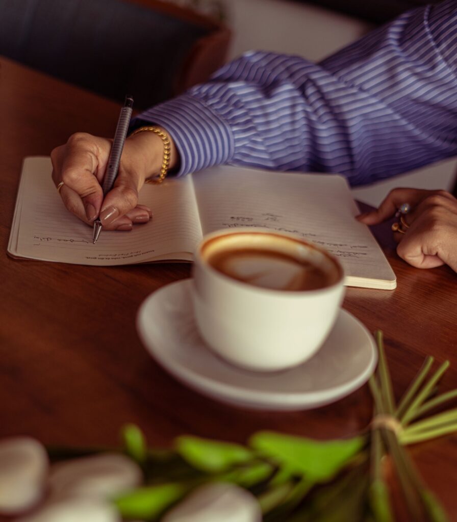A woman writes in a journal beside a cup of coffee and white tulips.