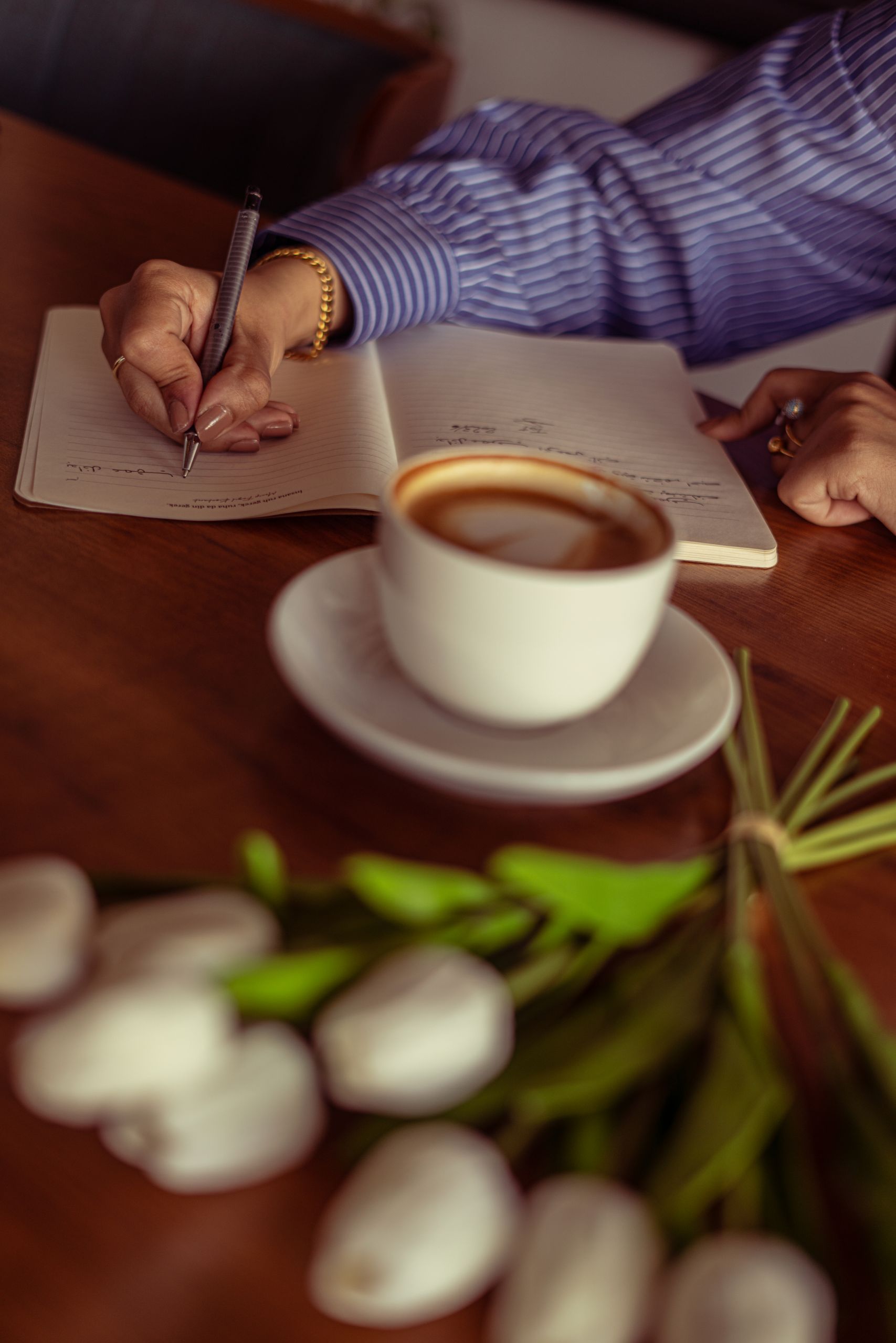 A woman writes in a journal beside a cup of coffee and white tulips.